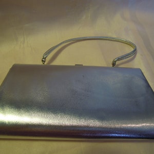 May include: A silver metallic clutch purse with a top handle. The purse has a smooth, shiny finish.