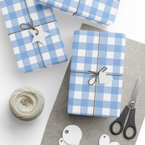 May include: Three rectangular packages wrapped in light blue and white gingham paper, tied with brown twine. Each package features a white paper gift tag in the shape of a star or heart. A ball of twine, scissors, and blank tags are present.