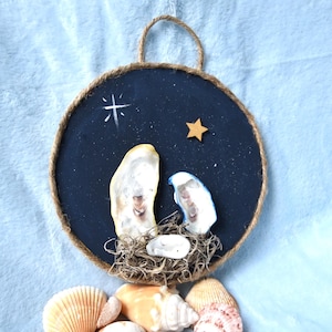 Può includere: Un presepe fatto a mano con conchiglie. L'opera d'arte circolare presenta uno sfondo blu scuro con una stella e una stella dorata. Le figure sono fatte di conchiglie e l'opera è rifinita con corda. Una collezione di conchiglie è in basso.