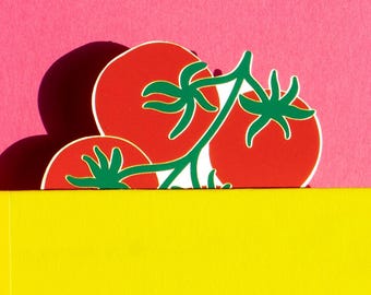 May include: A yellow book with a tomato-shaped bookmark. The bookmark features red tomatoes with green stems and leaves. The background is a solid pink color, creating a vibrant contrast.
