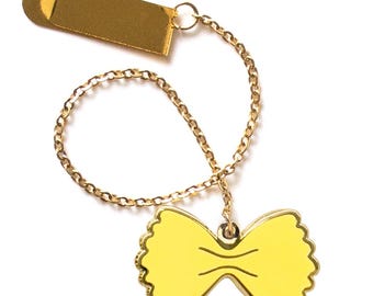 May include: A gold-colored pasta-shaped charm necklace. The charm is a yellow farfalle pasta shape with a gold outline, connected to a gold chain and a gold rectangular clasp. The necklace is set against a white background.
