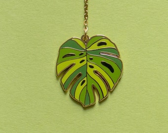 May include: A gold chain with a green and yellow monstera leaf charm. The leaf is outlined in gold.