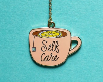 May include: A peach-colored enamel tea cup charm with a gold chain. The cup contains a lemon slice and the words "Self Care" in black script. A small tea bag with a heart is attached to the cup.