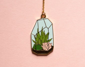 May include: A gold-colored necklace pendant featuring a terrarium design. The pendant has a light blue geometric frame with a black base, housing green succulents and a pink flower. The background is a solid light pink.