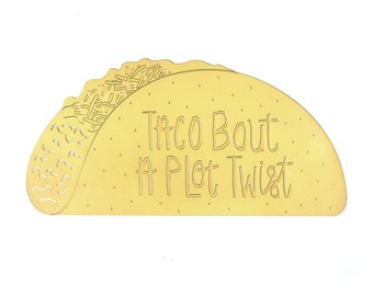 May include: A yellow taco-shaped sign with the text "Taco Bout a Plot Twist" written in a brown font.