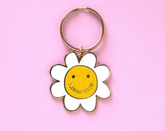 May include: A daisy-shaped keychain with a gold-colored ring. The flower has white petals and a yellow glittery center with a smiling face and the words "I joyfully decline". The keychain is on a pink background.