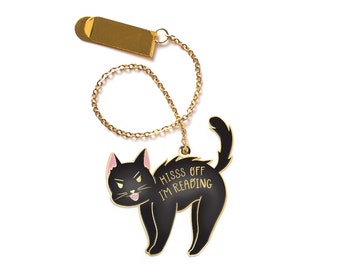 May include: Gold metal bookmark with a black cat design. The cat is arched with its back legs extended and its front paws on the ground. The cat has a pink tongue sticking out and its eyes are narrowed. The text "HISS OFF I'M READING" is written in gold on the cat's back.