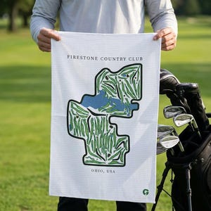 May include: A white waffle-textured golf towel featuring a printed map of the Firestone Country Club golf course in Ohio, USA. The design includes green fairways, blue water hazards, and black outlines, with the text Firestone Country Club and Ohio, USA.