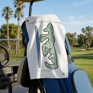 May include: A white waffle-textured golf towel featuring a green graphic illustration of a golf course layout, a blue vertical line, and the text "TORREY PINES GOLF COURSE" and "CALIFORNIA, USA" printed in black, with a small green logo at the bottom.