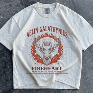 May include: An off-white short-sleeve t-shirt featuring a graphic of a deer with antlers and a crown surrounded by orange flames. The text reads AELIN GALATHYNIUS, FIREHEART, and includes two lines of small text beneath the deer illustration.