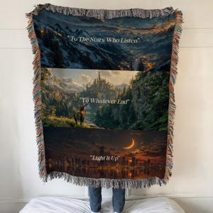 May include: Woven throw blanket featuring three landscape scenes with text: "To The Stars Who Listen," "To Whatever End," and "Light It Up." The blanket has a fringed edge and depicts mountains, a castle, a deer, and a cityscape.