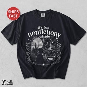 Pode incluir: Camiseta preta com uma ilustração fantástica. O design mostra uma pessoa olhando por uma janela para uma paisagem mágica, com o texto "it's too nonfictiony outside". A camiseta tem gola redonda e mangas curtas.