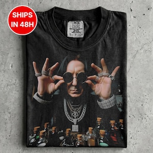 Puede incluir: Una camiseta negra con un estampado gráfico de una persona con cabello largo y oscuro, gafas de sol redondas, varias cadenas de plata y anillos en los dedos. La persona está situada detrás de varias botellas de vidrio pequeñas. La etiqueta interior dice COMFORT COLORS.