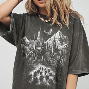 May include: A charcoal grey oversized t-shirt featuring a white graphic print of a large castle on a cliffside overlooking a lake with small boats carrying lanterns. A flying owl is depicted above the castle, set against a backdrop of mountains.
