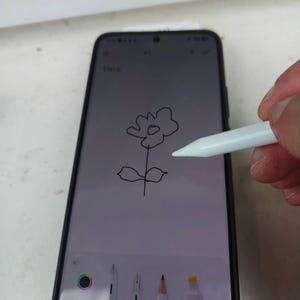 Peut inclure: Un smartphone noir avec un stylet blanc dessinant une fleur. L'écran affiche un croquis de fleur avec une tige. Le mot "Titre" est visible en haut. Le téléphone est dans un étui noir.