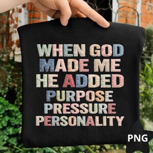 Può includere: Felpa nera con la scritta "WHEN GOD MADE ME HE ADDED PURPOSE PRESSURE PERSONALITY" in lettere colorate in stile patchwork. La felpa è tenuta in mano, mostrando il design.
