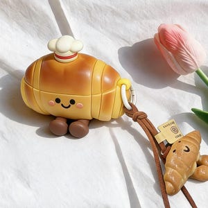 May include: A novelty croissant-shaped case in yellow and brown, complete with a chef's hat, smiling face, and brown feet. A small, brown leather strap holds a miniature croissant charm. A pink tulip is in the background.
