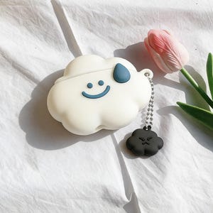 Puede incluir: Una funda blanca con forma de nube con una cara sonriente y un diseño de gota de lluvia azul. Un pequeño dije de nube negra con una cara enojada cuelga de una cadena. Sobre una superficie blanca con un tulipán rosa.