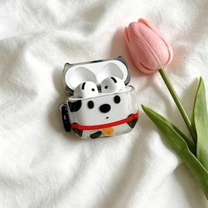 May include: A white and black spotted dog-themed protective case for wireless earbuds, featuring a red collar with a yellow tag, containing two white earbuds inside, positioned next to a single pink tulip flower with a green stem and leaves.