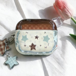May include: A clear case for wireless earbuds with a brown top and a star pattern. The case has a silver keychain with star-shaped charms. The stars are in shades of brown and blue.