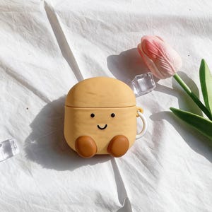 Puede incluir: Una funda para AirPods amarilla de estilo caricatura con forma de taza sonriente con pies marrones. La funda tiene un asa blanca y una parte superior redondeada. Un tulipán rosa y cubitos de hielo transparentes en el fondo.