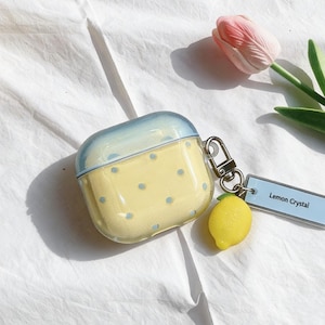 May include: A yellow and blue AirPod case with a lemon charm and a tag that reads "Lemon Crystal." The case has a yellow base with blue polka dots and a blue top. A silver clasp is attached to the case.