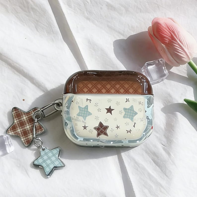 May include: A clear case for wireless earbuds with a brown top and a star pattern. The case has a silver clasp with two star-shaped charms, one plaid and one blue. A pink tulip and clear cubes are in the background.