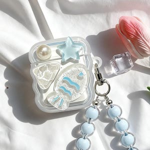 Puede incluir: Funda decorativa transparente para AirPod con diseño de pez, una estrella azul y una perla. La funda tiene una correa de cuentas azul claro y un cierre plateado. La funda está sobre una superficie blanca con un tulipán rosa y un cubo transparente.