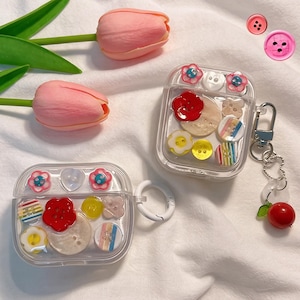 May include: Clear AirPod cases decorated with colorful buttons, flowers, and other embellishments. The cases have a keychain with a red cherry charm. Two pink tulips are in the background, along with a few loose buttons.