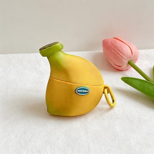 Op de afbeelding: Een gele banaanvormige hoes met een groene bovenkant en een klein blauw ovaal label met de tekst "BANANA". De hoes heeft een goudkleurige lus en staat naast een roze tulp.