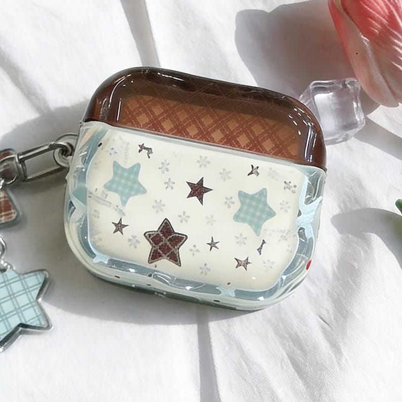 May include: Clear protective case for wireless earbuds. The case has a brown top with a plaid pattern and a white base decorated with blue and brown stars and snowflakes. A silver chain is attached to the case.
