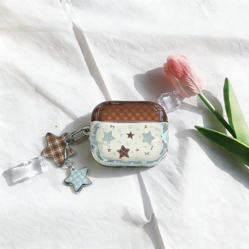 May include: A clear case for wireless earbuds with a brown top and a light blue and white patterned design. The case features a star pattern and a star-shaped charm. A pink tulip is in the background.