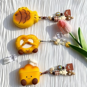 May include: Yellow silicone AirPod cases shaped like a fish, pretzel, and chef's hat. Each case has a matching charm with beads, stars, and other decorative elements. The cases are displayed on a white, textured surface.