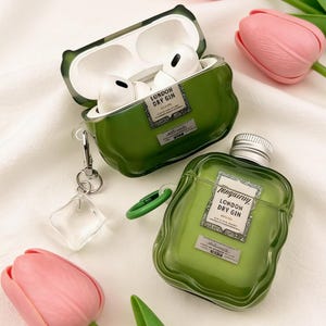 Peut inclure: Deux &eacute;tuis pour AirPods verts en forme de bouteilles de gin, avec l'inscription "London Dry Gin". L'un des &eacute;tuis est ouvert, r&eacute;v&eacute;lant des &eacute;couteurs blancs. Un charm transparent et une tulipe rose sont &eacute;galement visibles.