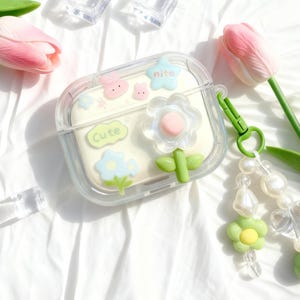 Puede incluir: Funda transparente para AirPod decorada con adornos de flores, conejos y estrellas en colores pastel. La funda tiene un clip verde y un charm de cuentas. La palabra "Cute" está escrita en la funda.
