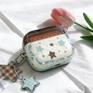 May include: A clear, protective case for wireless earbuds. The case has a brown top and a light blue base with a star pattern. A charm with two star-shaped pendants hangs from the side. A pink tulip and green leaves are in the background.