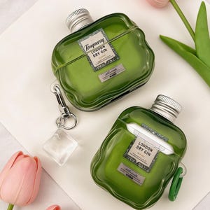 Peut inclure: Deux &eacute;tuis pour AirPod verts en forme de bouteille, avec des bouchons argent&eacute;s et l'inscription "Tanqueray London Dry Gin". L'un d'eux est muni d'un porte-cl&eacute;s argent&eacute; avec un charm carr&eacute; transparent. Une tulipe rose en arri&egrave;re-plan.