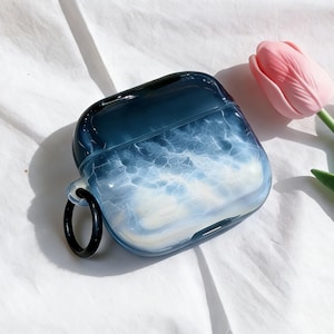 May include: A blue and white marbled AirPods case with a black ring attachment. The case has a glossy finish and a design that resembles clouds or ocean waves. A pink tulip is in the background.