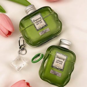 May include: Two green, bottle-shaped AirPod cases with silver tops and the "Tanqueray London Dry Gin" label. One case has a green loop and the other has a silver carabiner with a clear charm. Pink and green flowers are in the background.