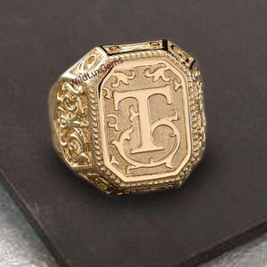 Op de afbeelding: Een goudkleurige zegelring met een gegraveerde letter "T" in een rechthoekig kader. De ring heeft sierlijke details aan de zijkanten en een gepolijste afwerking. De ring ligt op een donkere ondergrond.