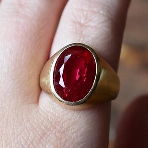 May include: A gold ring with a large, oval-cut, deep red gemstone. The ring has a simple, classic design with a smooth, polished band. The gemstone is the focal point, catching the light and displaying a vibrant color.