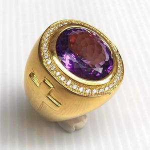May include: A gold ring with a large, oval purple gemstone. The gemstone is encircled by small, clear stones. The ring has a thick band with a cross-shaped design on the side.