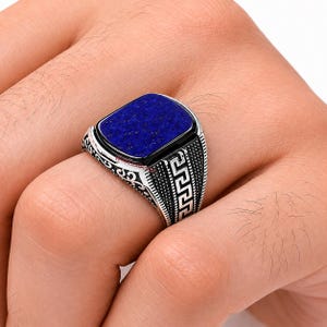 Könnte beinhalten: Ein silberner Ring mit einem rechteckigen, tiefblauen Stein. Der Ring hat einen schwarzen Rand um den Stein und ein detailliertes, gemustertes Design am Band. Der Ring befindet sich an einem Finger.