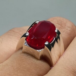 May include: A silver ring featuring a large, oval-cut, deep red gemstone. The ring has a modern, geometric design with a polished finish. The gemstone is the focal point, catching the light and showcasing its rich color.