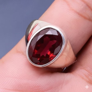 Könnte beinhalten: Ein silberner Ring mit einem großen, oval geschliffenen, tiefroten Edelstein. Der Edelstein ist in einem polierten Silberband gefasst. Der Ring wird an einem Finger gezeigt, um die Facetten und die Farbe des Edelsteins hervorzuheben.