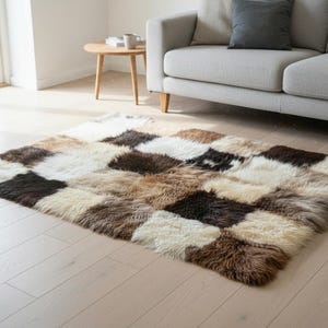 May include: A rectangular area rug with a patchwork design. The rug features squares of varying colors, including cream, brown, and black. The rug is made of a soft, textured material, likely wool or sheepskin, and is placed on a light-colored wooden floor.