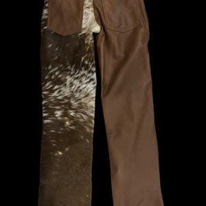 May include: Brown leather pants with a unique design. One leg features a cowhide pattern in shades of brown and white, while the other leg is solid brown leather. The pants have two back pockets and a classic five-pocket design.