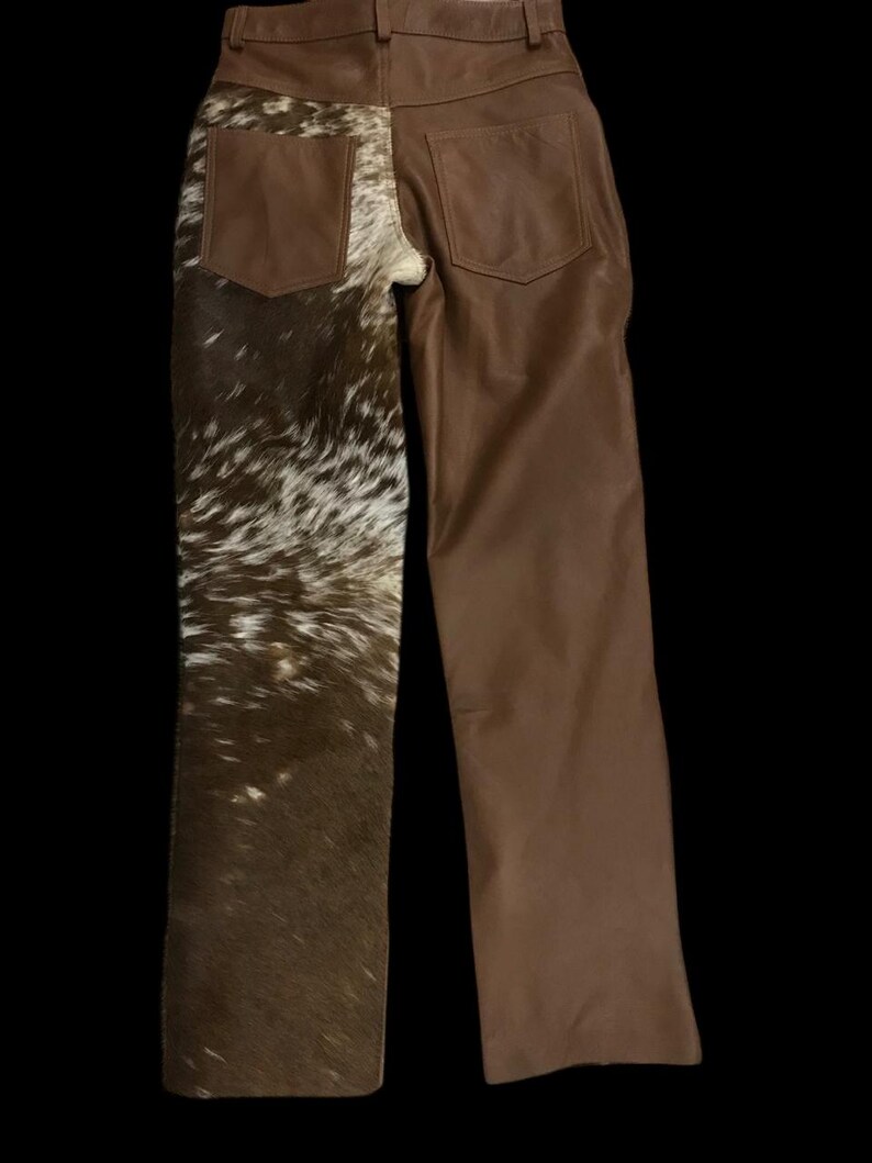 May include: Brown leather pants with a unique design. One leg features a cowhide pattern in shades of brown and white, while the other leg is solid brown leather. The pants have two back pockets and a classic five-pocket design.