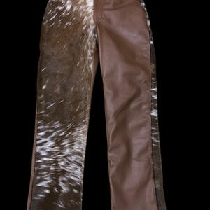 May include: Brown leather pants with a unique design. One leg features a cowhide pattern in shades of brown and white, while the other leg is solid brown leather. The pants have a classic five-pocket design.