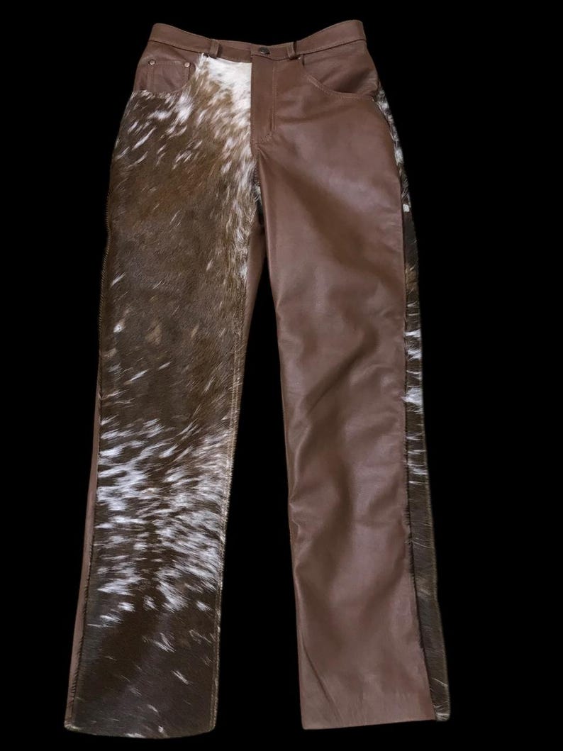 May include: Brown leather pants with a unique design. One leg features a cowhide pattern in shades of brown and white, while the other leg is solid brown leather. The pants have a classic five-pocket design.